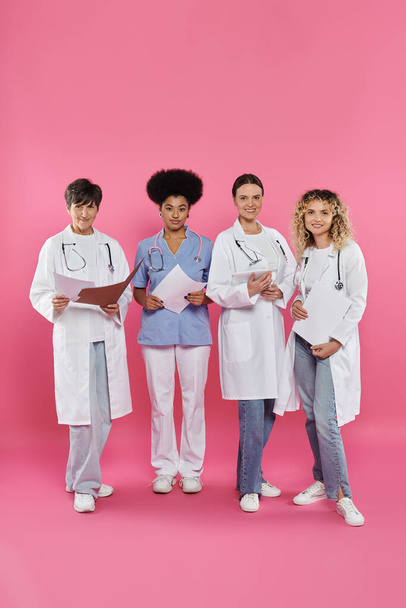 glimlachende interraciale artsen met papieren mappen en digitale tablet op roze, borstkanker maand - Foto, afbeelding