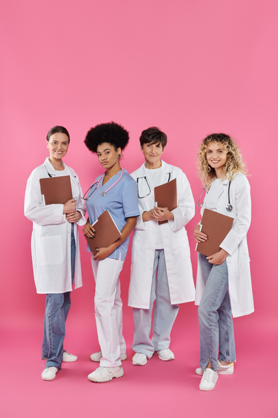 médicos multiétnicos sonrientes con carpetas de papel de pie juntos en rosa, conciencia del cáncer de mama - Foto, Imagen