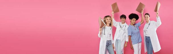 улыбающиеся многонациональные врачи с бумажными папками, изолированными на розовый, баннер, месяц рака молочной железы - Фото, изображение