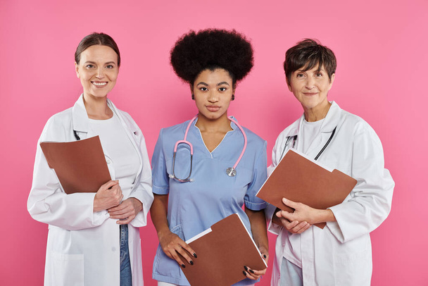 sonrientes médicos interraciales con batas blancas sosteniendo carpetas de papel aisladas en rosa, cáncer de mama - Foto, Imagen