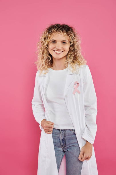 médico sorridente com fita no casaco branco de pé isolado no rosa, conceito de câncer de mama - Foto, Imagem
