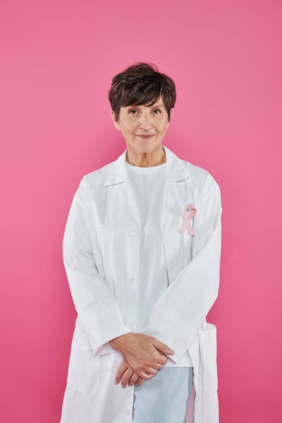 brunette doctor with ribbon on white coat standing isolated on pink, breast cancer concept - Photo, Image