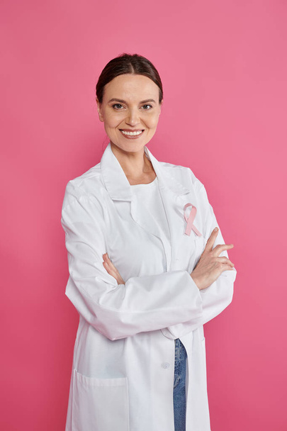 ピンクで隔離されたリボン交差腕を持つ陽気な女性腫瘍学者,乳癌月 - 写真・画像