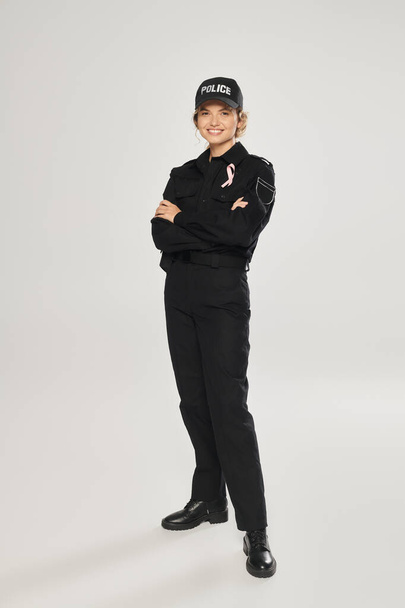 smiling policewoman with pink ribbon on uniform crossing arms on grey, breast cancer concept - Photo, Image