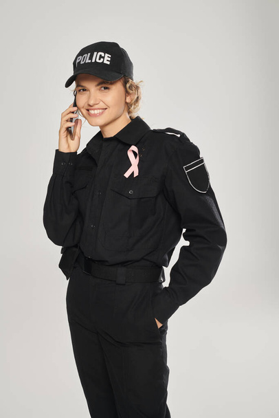 glückliche Polizistin mit rosafarbener Schleife an Uniform, die auf dem Smartphone mit grauer Isolierung spricht, Brustkrebs - Foto, Bild