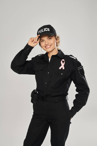 smiling policewoman with pink ribbon of breast cancer awareness looking at camera isolated on grey - Photo, Image