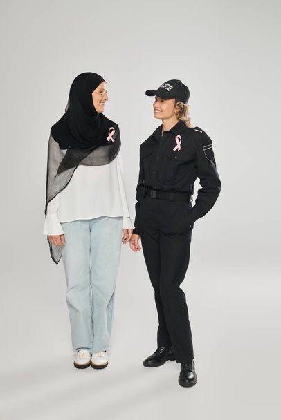 positive policewoman and woman in hijab with pink ribbons of breast cancer standing on grey - Foto, Imagem