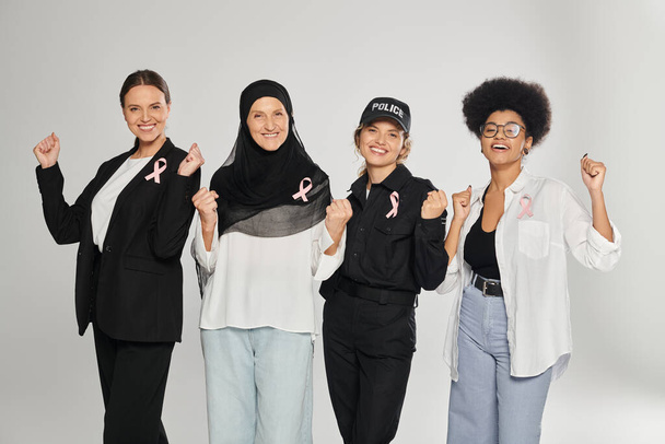 vzrušené a různé multietnické ženy s růžovými stuhami rakoviny prsu pózující izolované na šedé - Fotografie, Obrázek