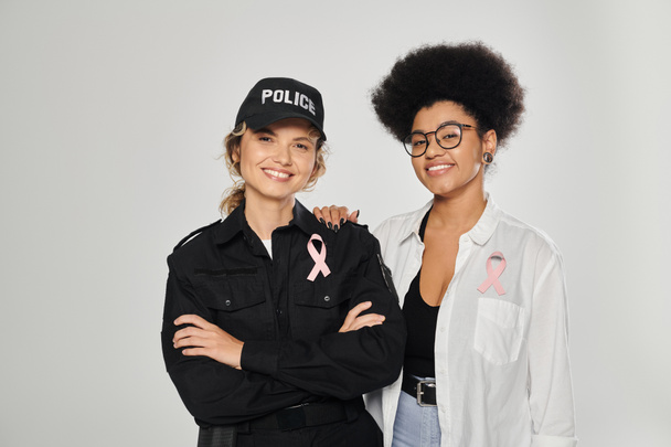 smiling african american woman with pink ribbon hugging policewoman isolated on grey, breast cancer - Photo, Image