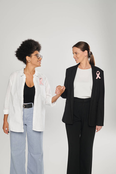 smiling businesswoman and african american woman with pink ribbons holding hands isolated on grey - 写真・画像