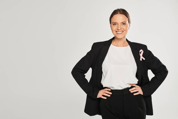 usmívající se podnikatelka s růžovou stuhou na bundě pózující na kameře izolované na šedé, rakovina prsu - Fotografie, Obrázek