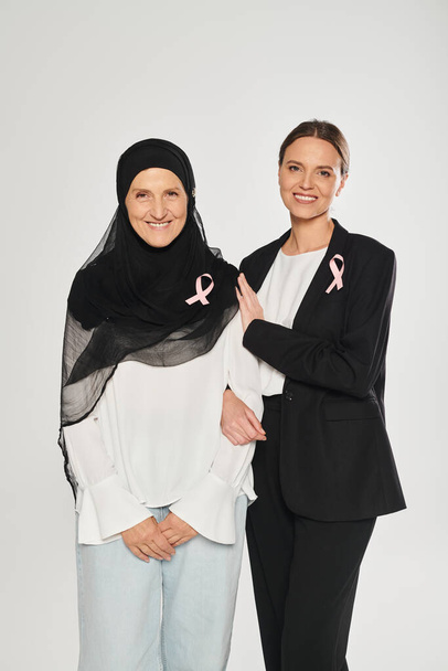 Улыбающаяся деловая женщина обнимает женщину в хиджабе с розовой лентой, изолированной на сером, месяц рака груди - Фото, изображение