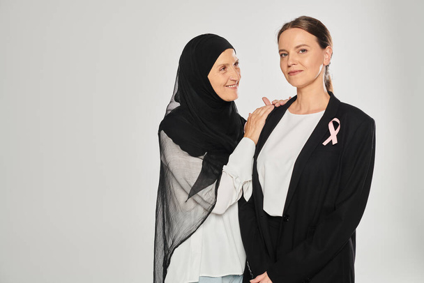 smiling woman in hijab hugging businesswoman with pink ribbon isolated on grey, breast cancer - Photo, image