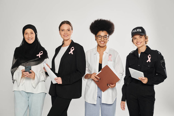 sorrindo diferentes mulheres inter-raciais com fitas rosa segurando dispositivos e papéis isolados em cinza - Foto, Imagem