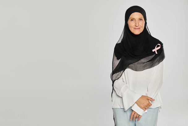 donna allegra in hijab e nastro rosa di consapevolezza del cancro al seno in piedi isolato su grigio - Foto, immagini