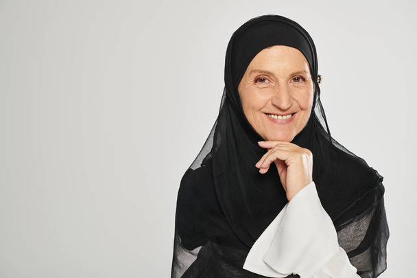 Porträt einer fröhlichen Frau im Hijab, die in die Kamera blickt, während sie isoliert auf grau posiert - Foto, Bild