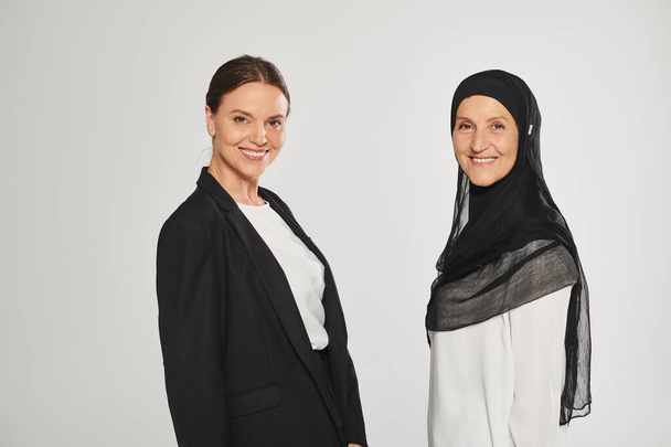 sonriente mujer de negocios y mujer en hijab mirando a la cámara juntos aislados en gris - Foto, imagen