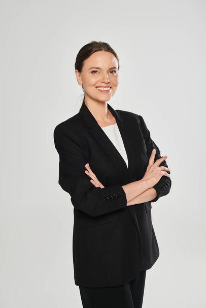 portrait of carefree woman in suit crossing arms while standing isolated on grey - Photo, Image