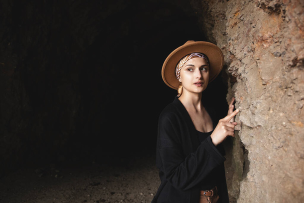 Piękna młoda kobieta w kapeluszu stoi przy ścianie jaskini w osadzie starożytnych ludzi. historyczna koncepcja turystyki. - Zdjęcie, obraz