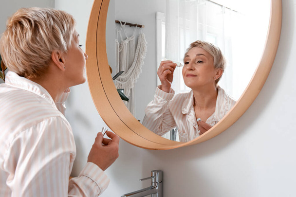 Mature woman applying under-eye cream near mirror in bathroom - Φωτογραφία, εικόνα