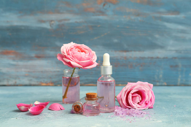 Бутылки косметического масла с экстрактом розы и цветами на синем столе - Фото, изображение