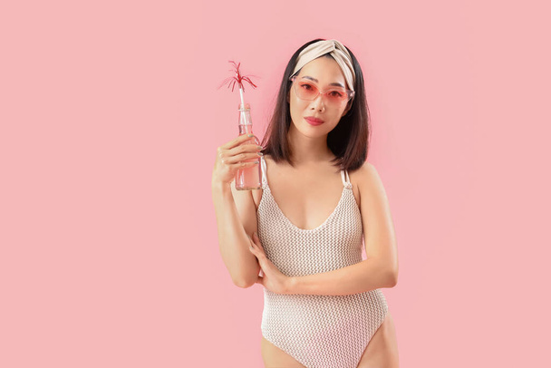 Beautiful Asian woman in swimsuit and with drink on pink background - Photo, Image