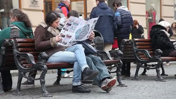 LVIV, UKRAINE - helmikuu 28, 2015 Kaksi turistia, mies ja nainen, opiskelu nähtävyyksiä kartta istuessaan penkillä ja ympäröivät kiireinen kaupunkielämä
 - Materiaali, video
