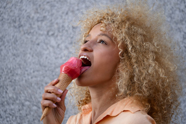 Gri arkaplanda izole edilmiş pembe dondurma yiyen gülümseyen mutlu bir kadının yan görüntüsü.. - Fotoğraf, Görsel