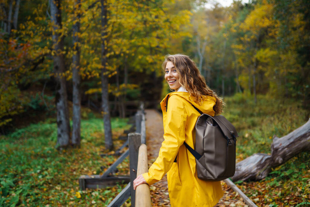 Autumn hike in nature. Woman in a yellow coat with a backpack walks along a wooden staircase, a path, enjoying the autumn scenery in the forest. Concept of nature, hiking, travel. - Foto, Imagen