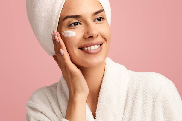 Positieve jonge vrouw in badjas verzorgen van haar huid, het aanbrengen van hydraterende crème op het gezicht, terwijl op roze achtergrond in de studio - Foto, afbeelding
