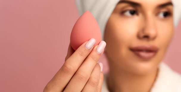 Soft focus of young woman in white bathrobe with towel on head showing makeup sponge to camera while looking away against pink background - Foto, Bild