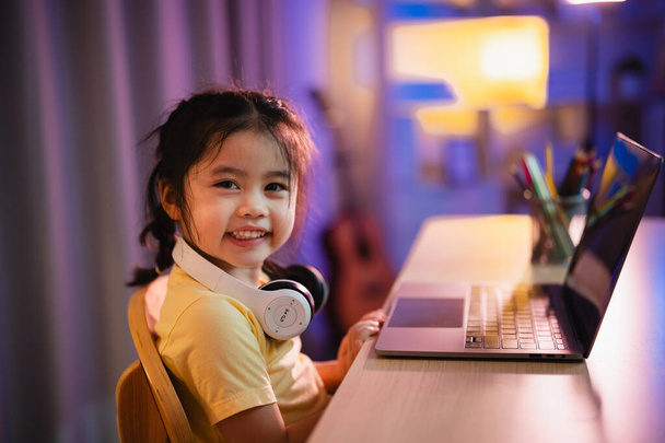 Aziatisch meisje glimlachen, lachen, opgewonden, kijken, het dragen van een witte hoofdtelefoon en het gebruik van laptop streaming nachtlampje in de woonkamer thuis. Schattig meisje kijken video 's terwijl tv Internet verslaving concept. - Foto, afbeelding