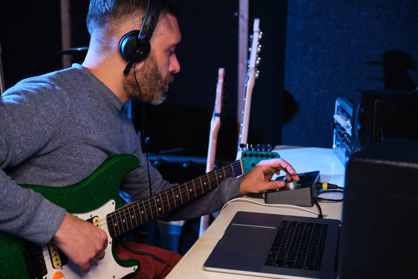 Muzikant man past het geluid van zijn elektrische gitaar met equalizer, koptelefoon en laptop thuis opnamestudio. Muziekproductie. - Foto, afbeelding