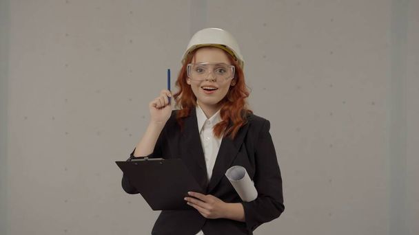 A woman in a helmet and with blueprints under her arm in the studio on a gray background. A woman architect in protective glasses happily exclaims there is an idea - Foto, imagen