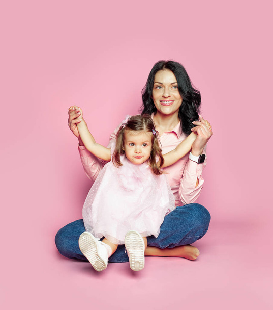 Feliz sonriente madre e hija sentadas sobre fondo de la pared del estudio rosa. Mamá y niño bebé niña estudio retrato - Foto, imagen