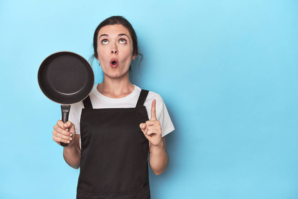 Woman with apron and pan on blue background pointing upside with opened mouth. - Photo, Image