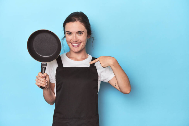 Femme avec tablier et casserole sur fond bleu personne pointant à la main vers un espace de copie chemise, fière et confiante - Photo, image