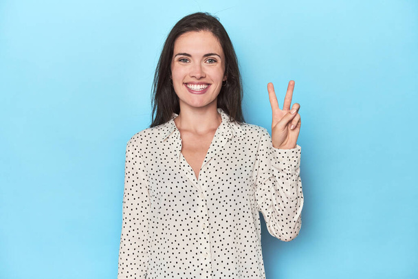 Young caucasian woman on blue backdrop showing number two with fingers. - Photo, Image
