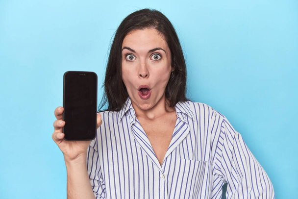 Удивлённая женщина показывает экран телефона на синем - Фото, изображение