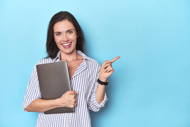 Jonge zakenvrouw houdt laptop op blauw lachend en wijzend opzij, toont iets op lege ruimte. - Foto, afbeelding