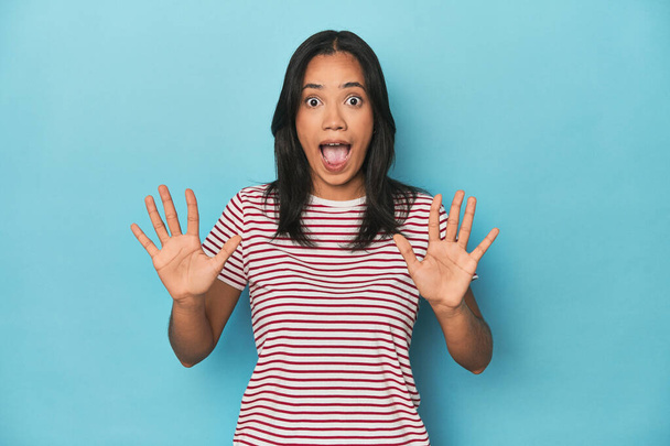 Φιλιππινέζα νεαρή γυναίκα στο μπλε στούντιο λαμβάνει μια ευχάριστη έκπληξη, ενθουσιασμένοι και σηκώνοντας τα χέρια. - Φωτογραφία, εικόνα