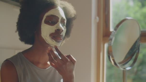 Střední zblízka záběr mladé afroamerické ženy aplikující hliněnou masku na pleť obličeje a při pohledu do stolního zrcadla během krásy rutiny doma - Záběry, video