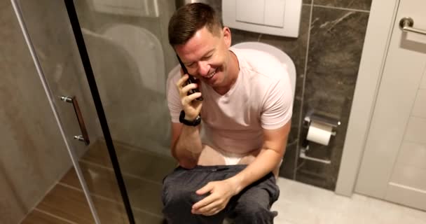 Le jeune homme parle au téléphone alors qu'il est assis sur les toilettes. Communication à distance dans les toilettes - Séquence, vidéo