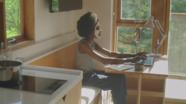 Střední dlouhý výstřel mladé Afroameričanky s hliněnou maskou na obličeji pracuje na notebooku v kuchyni doma - Záběry, video