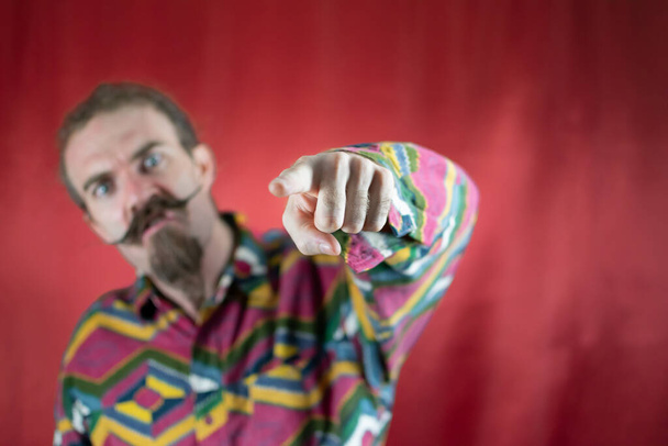 Hombre hipster enojado apuntando a la cámara gritando y con expresión furiosa con fondo rojo - Foto, Imagen