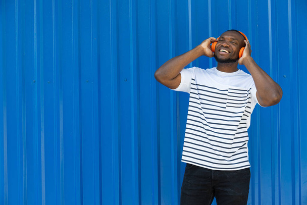 Un hombre afroamericano joven y elegante escucha música con auriculares sobre un fondo azul - Foto, Imagen