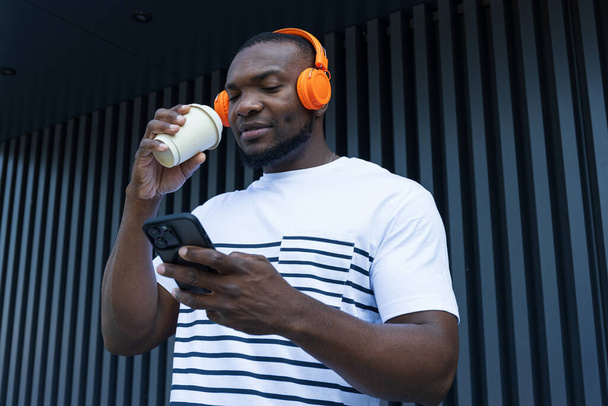 スタイリッシュで若いアフリカ系アメリカ人男性がヘッドフォンでコーヒーを手に入れて音楽を聴く. - 写真・画像