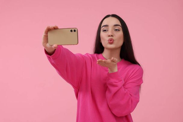 Young woman taking selfie with smartphone and blowing kiss on pink background - 写真・画像