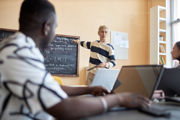 Focus on young confident man in casualwear pointing at formulas on blackboard while explaining them to group of classmates at lesson - Photo, Image