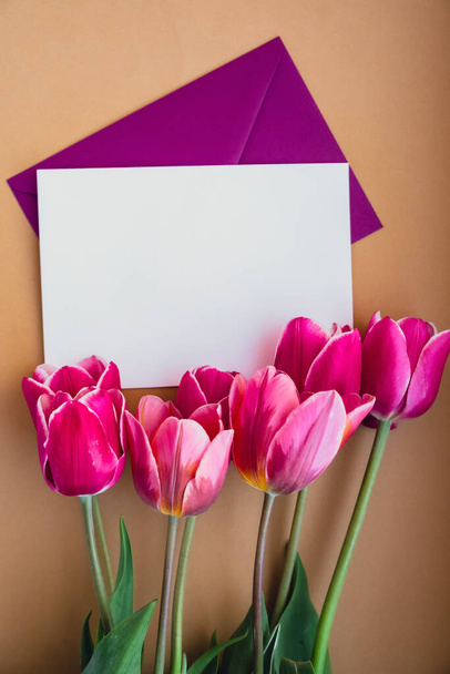 Empty white envelope copy space for your text or design with beautiful pink tulip bouquet on beige background. Template mock up for holiday spring greeting card. Top view Flat lay blank paper Feminine - Photo, Image